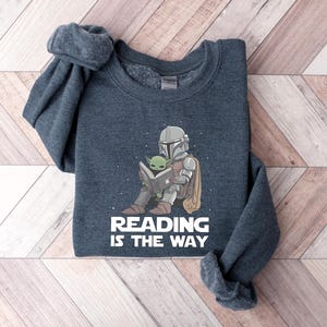 May include: Dark gray sweatshirt featuring a cartoon illustration of a Mandalorian character and Grogu reading a book. The text "READING IS THE WAY" is printed in white below the illustration. The sweatshirt has a crew neck and long sleeves.