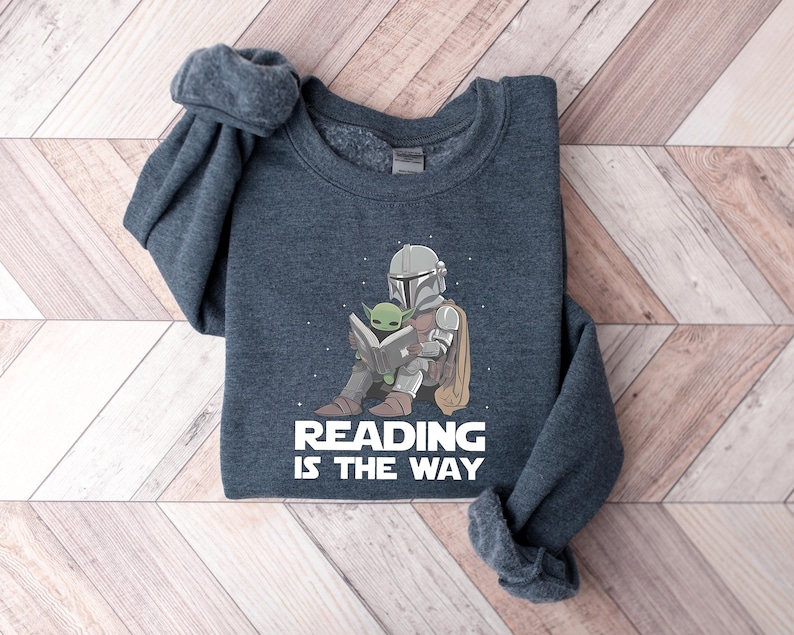 May include: Dark gray sweatshirt featuring a cartoon illustration of a Mandalorian character and Grogu reading a book. The text "READING IS THE WAY" is printed in white below the illustration. The sweatshirt has a crew neck and long sleeves.