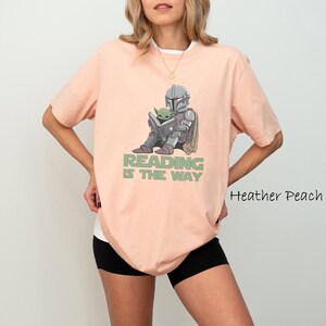 May include: Heather peach-colored t-shirt with a graphic of a character in armor reading a book with a small green creature. The text "READING IS THE WAY" is printed below the graphic. The shirt has short sleeves and a white undershirt.