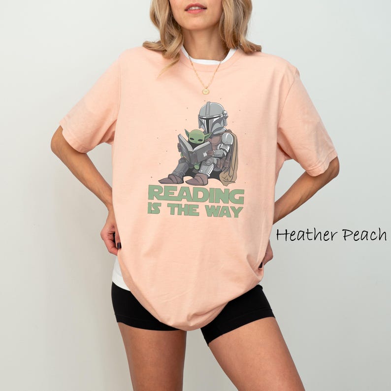 May include: Heather peach-colored t-shirt with a graphic of a character in armor reading a book with a small green creature. The text "READING IS THE WAY" is printed below the graphic. The shirt has short sleeves and a white undershirt.