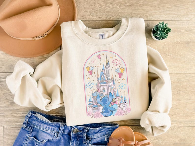 K&ouml;nnte beinhalten: Cremefarbenes Sweatshirt mit einem skurrilen Schlossdesign mit Feuerwerk, Luftballons und einer Cartoonfigur. Das Design ist von einem rosa Bogen und floralen Akzenten umrahmt. Das Sweatshirt wird mit blauen Jeansshorts und Sandalen kombiniert.