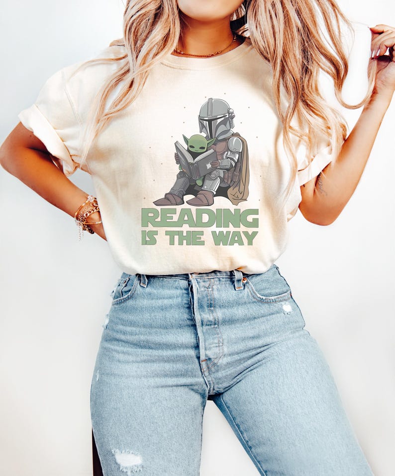 May include: Cream-colored t-shirt featuring a graphic of a Mandalorian character and Grogu reading a book. The text "READING IS THE WAY" is printed in green below the illustration. The shirt is paired with light blue denim jeans.