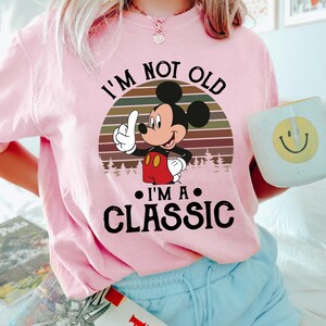 Puede incluir: Camiseta rosa claro con Mickey Mouse y el texto "I'm Not Old, I'm a Classic." El dise&ntilde;o incluye un atardecer retro y una silueta de bosque. Se complementa con pantalones cortos azul claro y una taza con una carita sonriente.