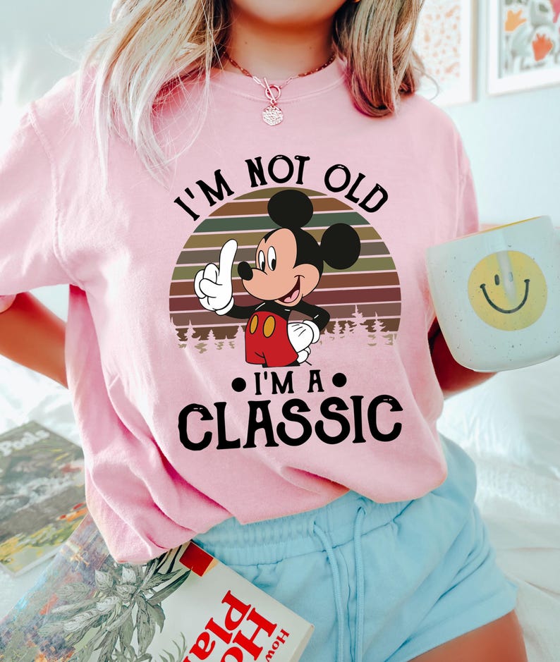 Puede incluir: Camiseta rosa claro con Mickey Mouse y el texto "I'm Not Old, I'm a Classic." El dise&ntilde;o incluye un atardecer retro y una silueta de bosque. Se complementa con pantalones cortos azul claro y una taza con una carita sonriente.