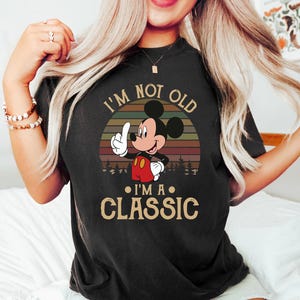 Puede incluir: Camiseta negra con Mickey Mouse y el texto "I'm Not Old, I'm a Classic." El dise&ntilde;o incluye un atardecer retro y una silueta de bosque. Mickey se&ntilde;ala hacia arriba, con pantalones cortos rojos y guantes blancos.