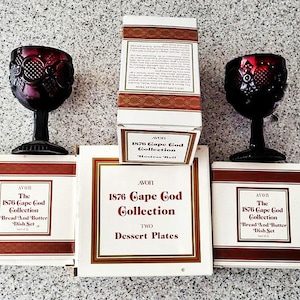 May include: A display of Avon's 1876 Cape Cod Collection glassware and dish sets. The image shows two ruby-coloured goblets and several boxes with the collection's branding. The boxes contain bread and butter dish sets and dessert plates.