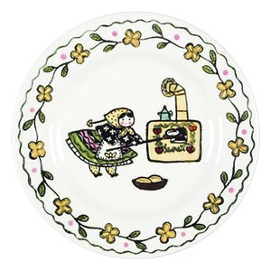 Könnte beinhalten: Ein weißer Keramikteller mit einem grünen, gelben und rosafarbenen Blumensaum. In der Mitte des Tellers befindet sich eine handgemalte Illustration einer Frau in einem grün-rosa-weißen Kleid, die einen Topf auf einem Herd umrührt.