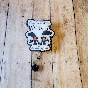 May include: A badge reel with the text "There is a little Witch in all of us." The badge features two witch figures holding umbrellas. The reel has a black glitter button and a clear plastic retractable cord.