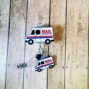 May include: Two white and red mail truck badge reels. The badge reels have a clear retractable cord and a metal clip. The mail trucks have the word "MAIL" in red letters. The background is a light brown wood.