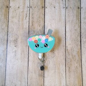 May include: A teal badge reel featuring a cartoon bowl of cereal with a smiling face. The cereal pieces are colorful, and the reel has a silver glitter rim and a silver spoon. The reel has a retractable cord and a black glitter button.