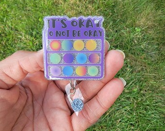 May include: A rectangular badge reel with the text "IT'S OKAY TO NOT BE OKAY" in purple. The badge has a gradient of colorful circles and a glittery border. The reel is held in a hand against a green grass background.