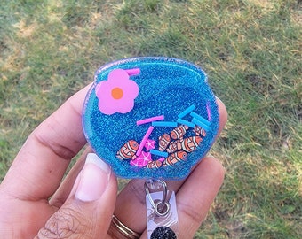 May include: A blue glitter resin keychain with a pink flower, orange center, and orange and white fish. The keychain has a clear plastic badge holder with a black glitter circle.