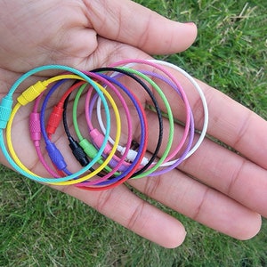 May include: A collection of colorful metal key rings in various colors, including yellow, pink, blue, green, and black. The key rings are made of braided metal cable and have a metal clasp.