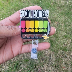 May include: A silver glitter badge holder with a black and white battery graphic that says "SOCIAL BATTERY". The battery has a rainbow color bar with six different colored sections. Each section has a different emoji face, starting with a sad face and ending with a happy face. The badge holder has a clear plastic retractable badge holder with a blue glitter circle.