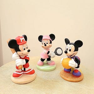 May include: Three ceramic figurines of Mickey Mouse and Minnie Mouse playing different sports. Mickey Mouse is wearing a red baseball uniform and holding a bat, Minnie Mouse is wearing a pink tennis outfit and holding a tennis racket, and Mickey Mouse is wearing a purple basketball uniform and holding a basketball.