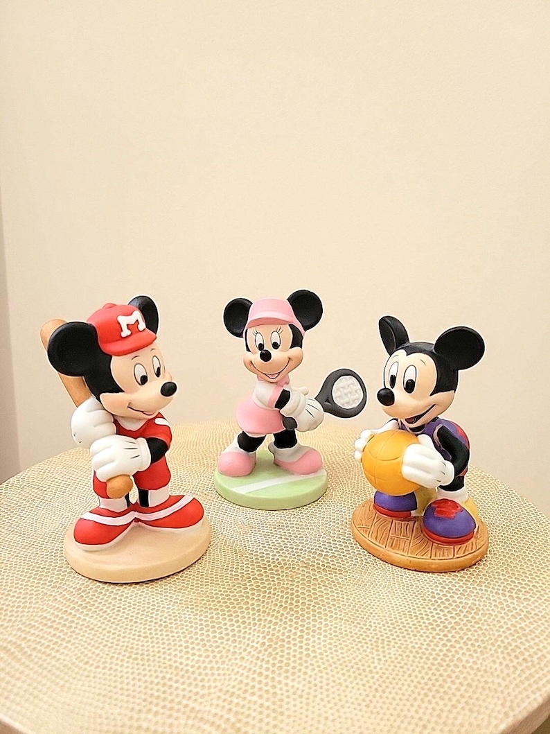 May include: Three ceramic figurines of Mickey Mouse and Minnie Mouse playing different sports. Mickey Mouse is wearing a red baseball uniform and holding a bat, Minnie Mouse is wearing a pink tennis outfit and holding a tennis racket, and Mickey Mouse is wearing a purple basketball uniform and holding a basketball.