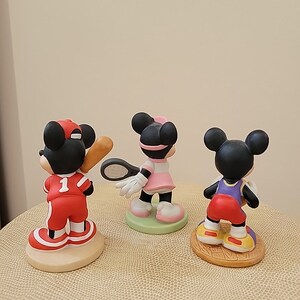 May include: Three ceramic figurines of Mickey Mouse dressed in different sports uniforms. The first figurine is wearing a red baseball uniform with a baseball bat. The second figurine is wearing a pink tennis uniform with a tennis racket. The third figurine is wearing a purple basketball uniform with a basketball.