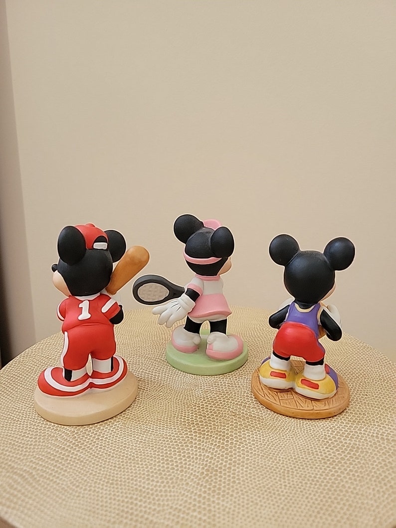 May include: Three ceramic figurines of Mickey Mouse dressed in different sports uniforms. The first figurine is wearing a red baseball uniform with a baseball bat. The second figurine is wearing a pink tennis uniform with a tennis racket. The third figurine is wearing a purple basketball uniform with a basketball.