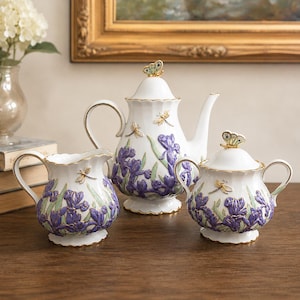 May include: A white porcelain tea set featuring a teapot, creamer, and sugar bowl. The set is decorated with purple iris flowers, green leaves, and gold accents. Each piece has a butterfly or dragonfly on the lid or body.
