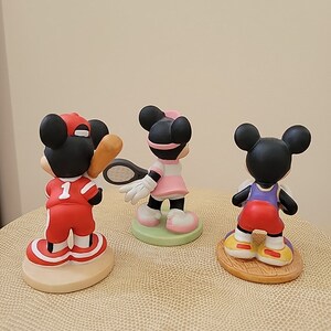 May include: Three ceramic figurines of Mickey Mouse dressed in different sports uniforms. The first figurine is wearing a red baseball uniform with the number 1 on the front. The second figurine is wearing a pink tennis dress and holding a tennis racket. The third figurine is wearing a red basketball uniform.