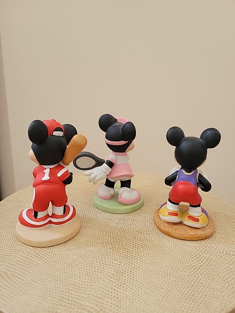 May include: Three ceramic figurines of Mickey Mouse dressed in different sports uniforms. The first figurine is wearing a red baseball uniform with the number 1 on the front. The second figurine is wearing a pink tennis dress and holding a tennis racket. The third figurine is wearing a red basketball uniform.