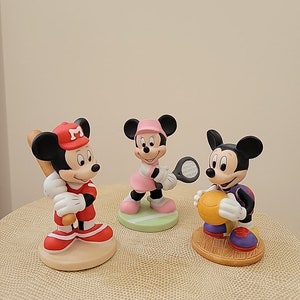 May include: Three ceramic figurines of Mickey Mouse characters playing different sports. The first figurine is Mickey Mouse wearing a red baseball uniform and holding a bat. The second figurine is Minnie Mouse wearing a pink tennis outfit and holding a tennis racket. The third figurine is Mickey Mouse wearing a purple basketball uniform and holding a basketball.