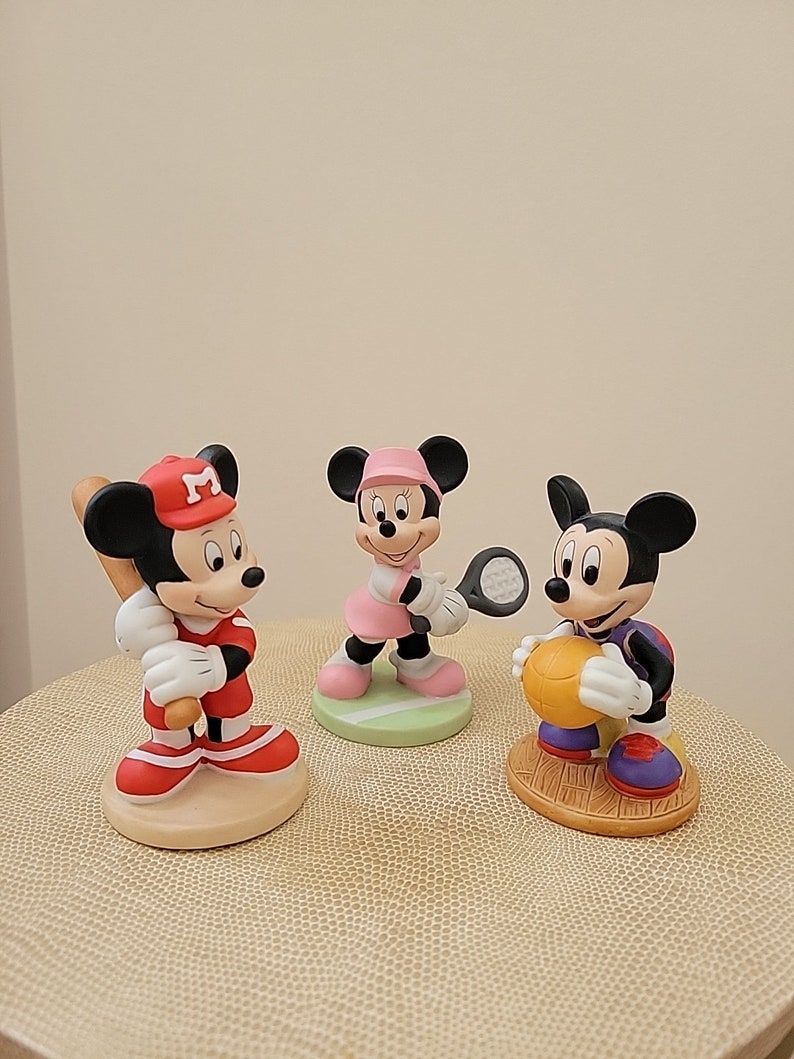 May include: Three ceramic figurines of Mickey Mouse characters playing different sports. The first figurine is Mickey Mouse wearing a red baseball uniform and holding a bat. The second figurine is Minnie Mouse wearing a pink tennis outfit and holding a tennis racket. The third figurine is Mickey Mouse wearing a purple basketball uniform and holding a basketball.