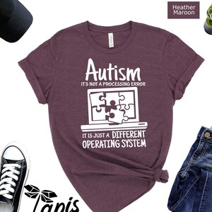 May include: A heather maroon t-shirt with a white graphic design that reads "Autism It's not a processing error It is just a different operating system". The graphic features a computer screen with a puzzle piece inside.