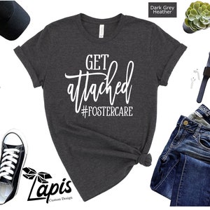 May include: Dark grey heather t-shirt with the text "GET attached #FOSTERCARE" in white lettering. The shirt is shown on a white background with other items like a hat, sunglasses, and jeans.