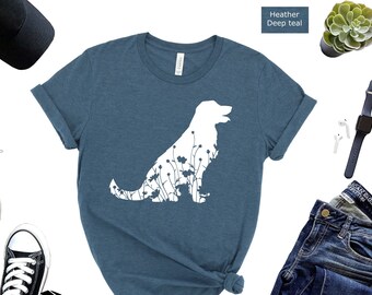 May include: A heather deep teal t-shirt with a white silhouette of a dog with flowers inside the outline. The dog is facing right and has its tail curled up. The t-shirt is laid flat on a white background with other items like a pair of black sneakers, sunglasses, a black baseball cap, a pair of blue jeans, a potted succulent, and a pair of white earbuds.