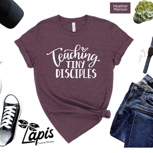 May include: Heather maroon t-shirt with the text "Teaching Tiny Disciples" in white lettering. The shirt is shown on a white background with a pair of black sneakers, a black baseball cap, and a pair of blue jeans.