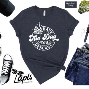 May include: A heather navy blue t-shirt with a white graphic that says "Have the Day You Deserve" with a skeleton hand giving a thumbs up.