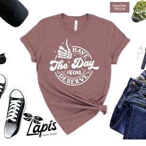 May include: A heather mauve t-shirt with a white graphic that says "Have the Day You Deserve" with a skeleton hand giving a thumbs up.