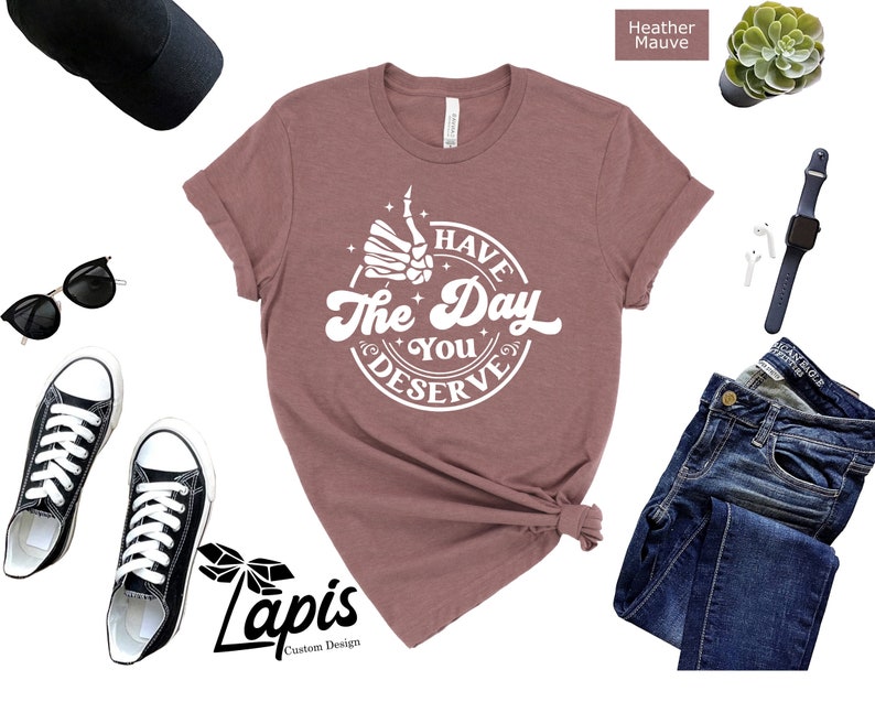 May include: A heather mauve t-shirt with a white graphic that says "Have the Day You Deserve" with a skeleton hand giving a thumbs up.