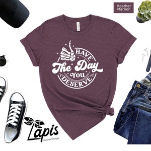 May include: A heather maroon t-shirt with a white graphic that says "Have the Day You Deserve" with a skeleton hand giving a thumbs up.
