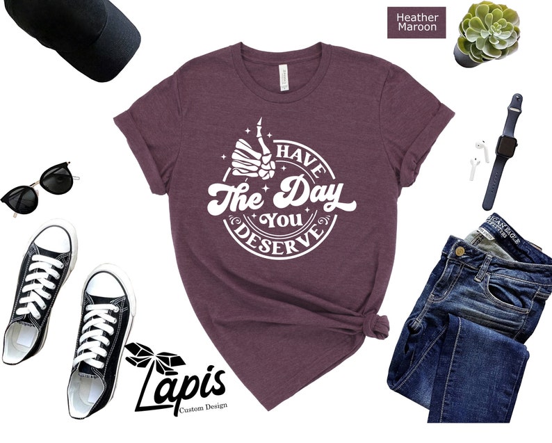May include: A heather maroon t-shirt with a white graphic that says "Have the Day You Deserve" with a skeleton hand giving a thumbs up.