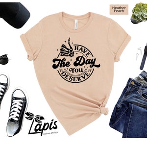 May include: A heather peach t-shirt with a black graphic that says "Have the Day You Deserve" with a skeleton hand giving a thumbs up.