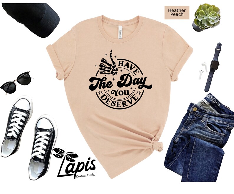 May include: A heather peach t-shirt with a black graphic that says "Have the Day You Deserve" with a skeleton hand giving a thumbs up.