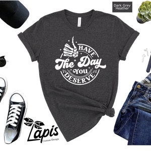 May include: Dark grey heather t-shirt with a white graphic of a skeleton hand giving a thumbs up with the text "Have the Day You Deserve".
