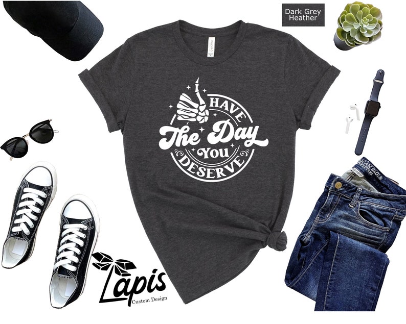 May include: Dark grey heather t-shirt with a white graphic of a skeleton hand giving a thumbs up with the text "Have the Day You Deserve".