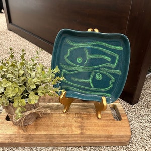 May include: A teal ceramic serving dish with a gold metal stand. The dish has a green design of two stylized fish.