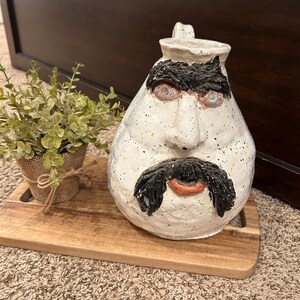 May include: White ceramic pitcher with a face painted on it. The face has black eyebrows, a black mustache, and a red mouth. The pitcher has a speckled white glaze with blue, yellow, and green accents.