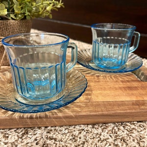 May include: Two light blue glass teacups with matching saucers. The cups have a ribbed design and are sitting on a wooden surface.