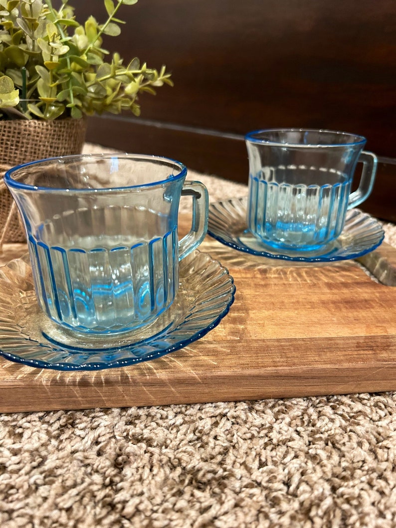 May include: Two light blue glass teacups with matching saucers. The cups have a ribbed design and are sitting on a wooden surface.
