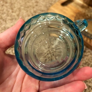 May include: A small, light blue glass cup with a textured rim. The cup is empty and appears to be made of clear glass.