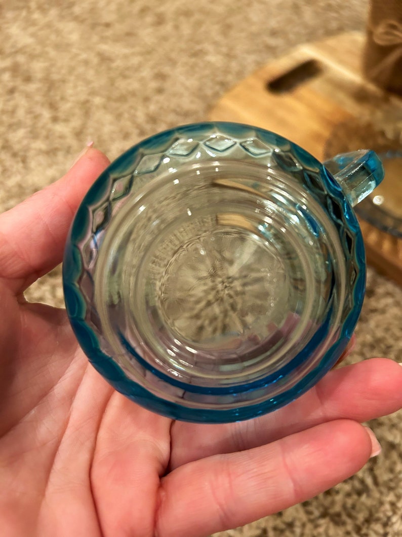 May include: A small, light blue glass cup with a textured rim. The cup is empty and appears to be made of clear glass.