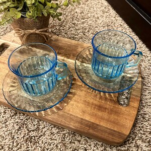 May include: Two blue glass teacups with matching saucers on a wooden serving tray. The cups have a ribbed design and a blue rim.