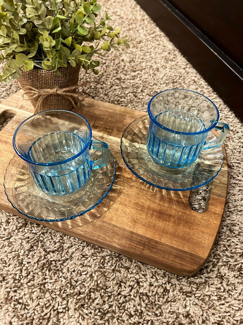May include: Two blue glass teacups with matching saucers on a wooden serving tray. The cups have a ribbed design and a blue rim.