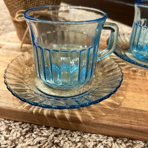 May include: Two blue glass teacups with matching saucers. The cups have a ribbed design and are sitting on a wooden surface.