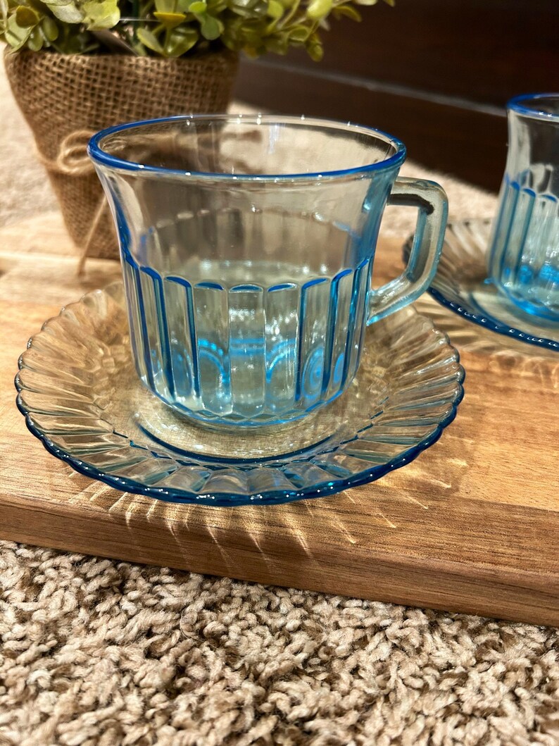 May include: Two blue glass teacups with matching saucers. The cups have a ribbed design and are sitting on a wooden surface.